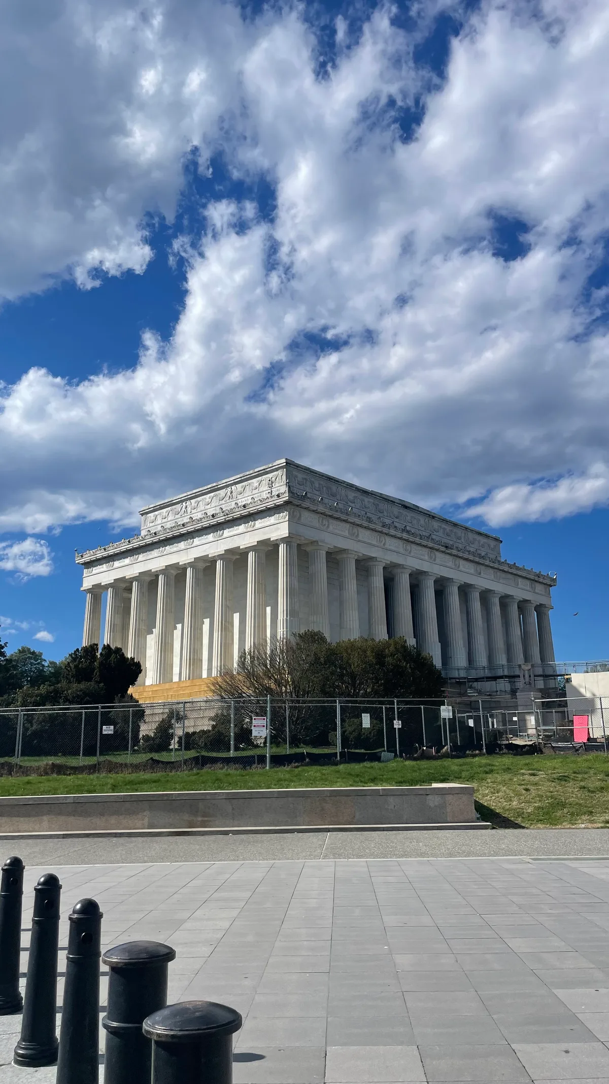 The Lincoln Memorial rises in bright white marble beneath a vivid blue sky streaked with dramatic, billowing clouds. Strong sunlight highlights the tall Doric columns, with a paved foreground, low black bollards, and a few trees framing the monument.