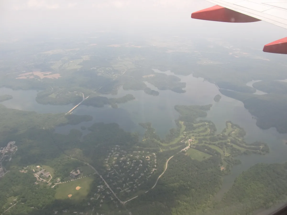 High, hazy aerial view of Loch Raven Reservoir, with calm gray water winding among forested peninsulas, scattered homes, and patches of farmland below. Soft morning mist and pale golden light flatten the distant horizon, giving the landscape a muted, atmospheric feel.