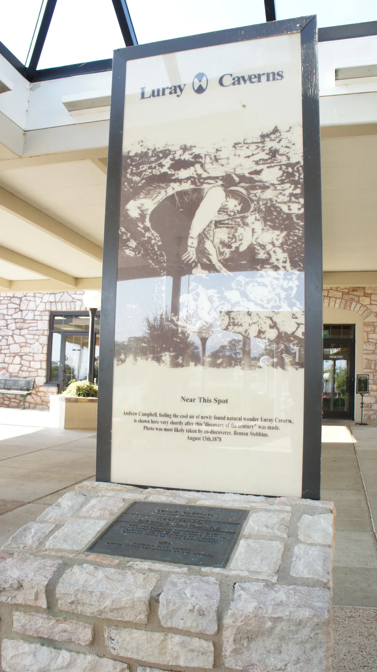 A tall outdoor sign at Luray Caverns stands in a stone base, showing a sepia-toned historic cave photo and the words “Luray Caverns” and “Near This Spot.” The covered entrance behind it has pale stone walls, dark window trim, and bright daylight filtering through the open roof.