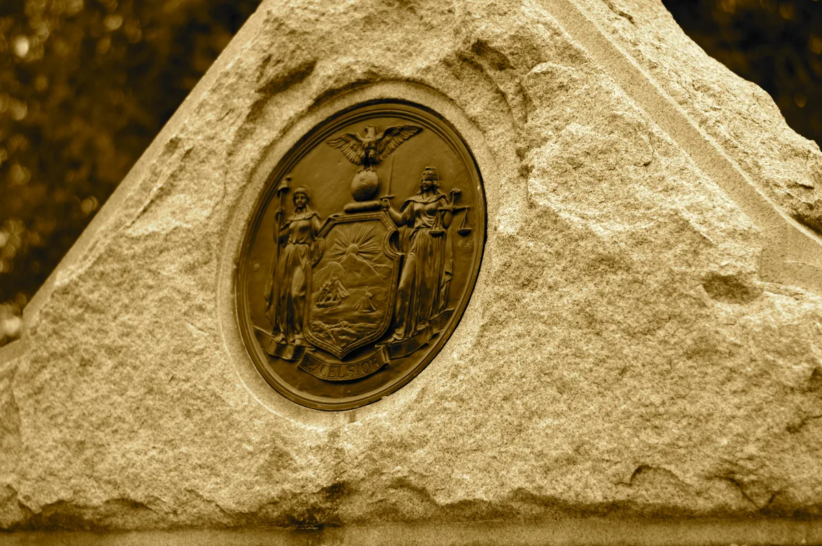 A close-up of a rough-hewn stone monument set in warm golden light, with a bronze Virginia state seal inset at the center showing the standing figure of Virtus. The background falls into a dark blur, making the textured granite and detailed medallion the focus.