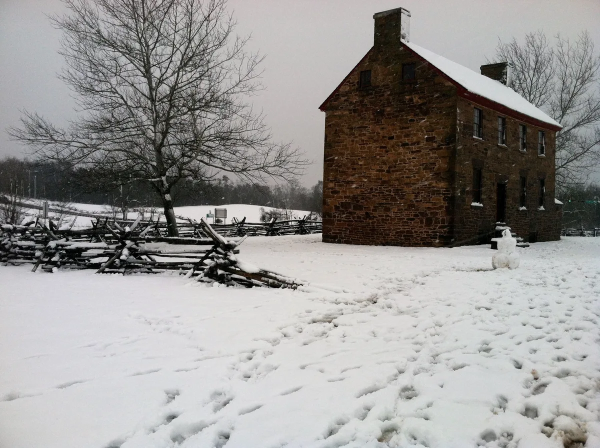 Snow blankets the battlefield around a dark stone hospital building, with a leafless tree and split-rail fence cutting across the foreground. Footprints mark the white ground under a gray winter sky, giving the scene a quiet, historic mood.