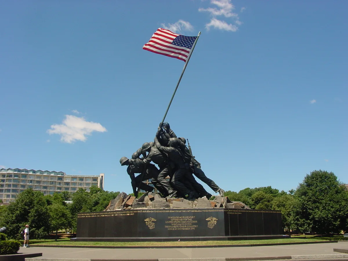 The Marine Corps War Memorial shows the bronze Iwo Jima flag-raising sculpture on a dark pedestal, with the American flag leaning high against a clear blue sky. Trees and a nearby building frame the monument in bright daylight.