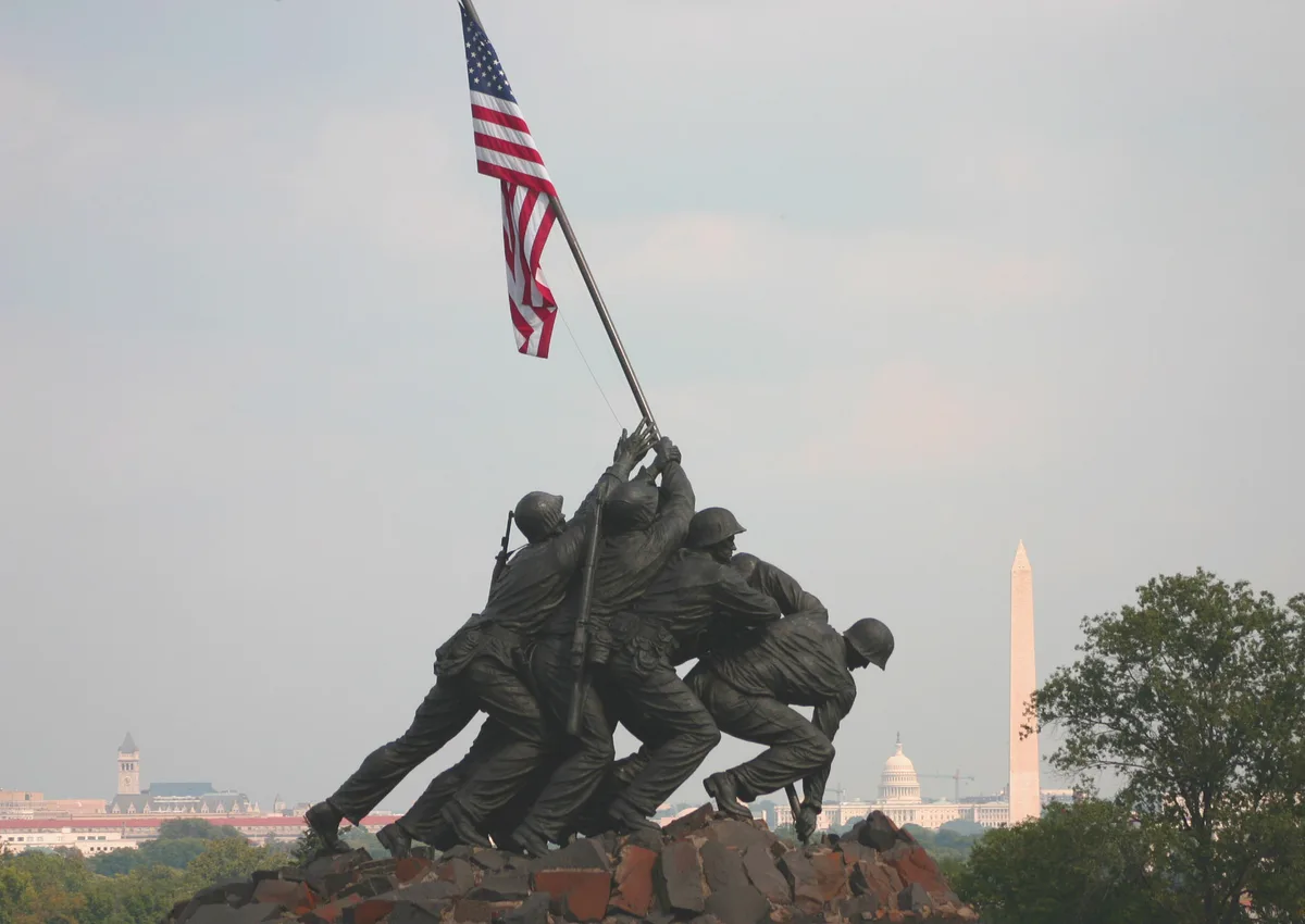 The bronze Marine Corps War Memorial shows the Iwo Jima flag-raising scene silhouetted against a pale sky, with the American flag lifted high above the figures. In the distance, the Washington Monument and U.S. Capitol dome rise over a line of trees, giving the monument a sweeping DC skyline backdrop.