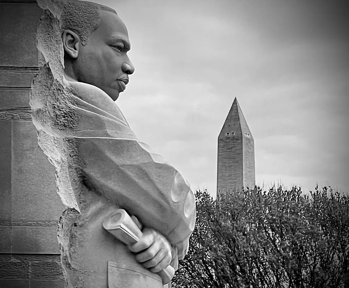 Black-and-white close-up of the Martin Luther King Jr. statue carved from the Stone of Hope, shown in profile with his folded arms in the foreground. The Washington Monument rises in the background above a line of trees, creating a strong layered composition.