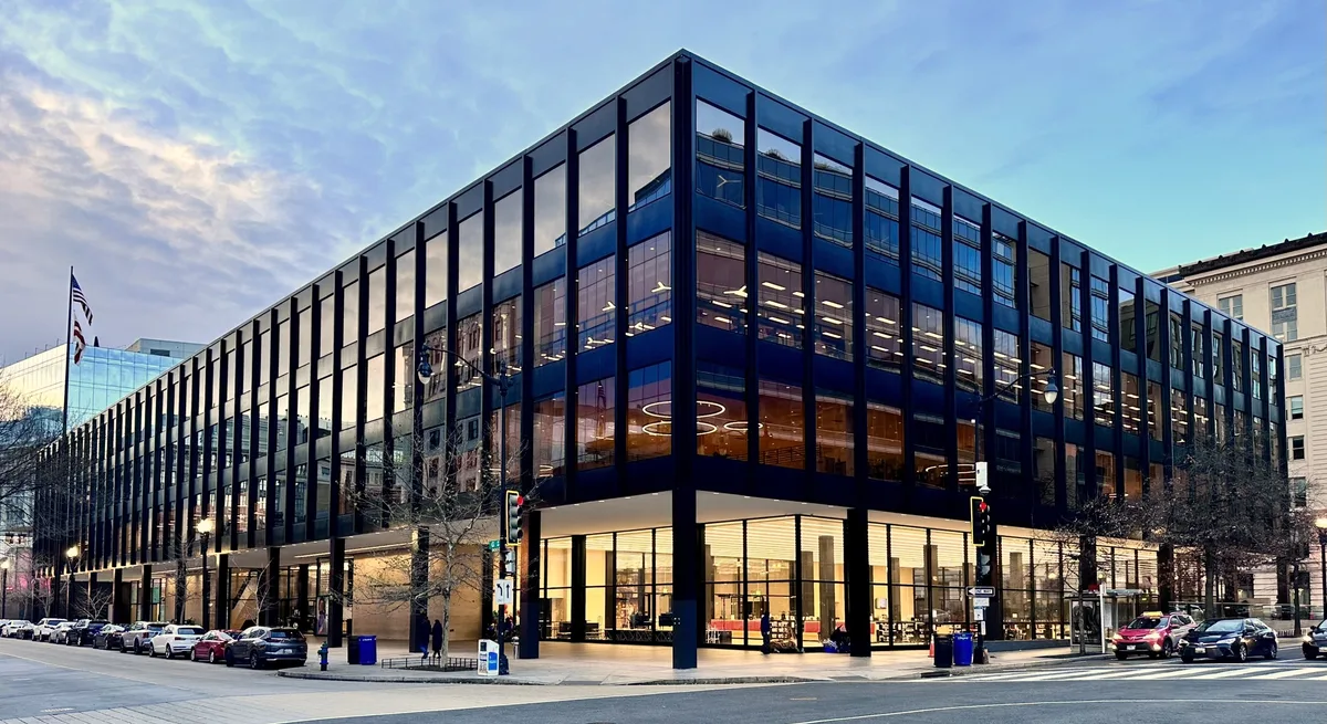 The Martin Luther King Jr. Memorial Library rises as a sleek black glass-and-steel cube, its grid of windows reflecting the blue evening sky and nearby buildings. Warm interior lights glow through the lower floors while cars line the street at the corner beneath a soft golden-hour cloud cover.