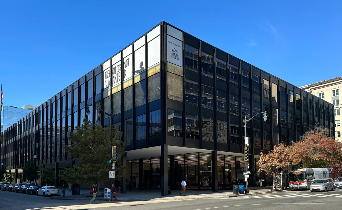 The Martin Luther King Jr. Memorial Library rises at a sharp corner in downtown Washington, its dark glass facade reflecting the street and bright blue sky. A large “Free to be is not equal to freedom” banner and the library’s long horizontal lines give the modern building a bold, graphic look.