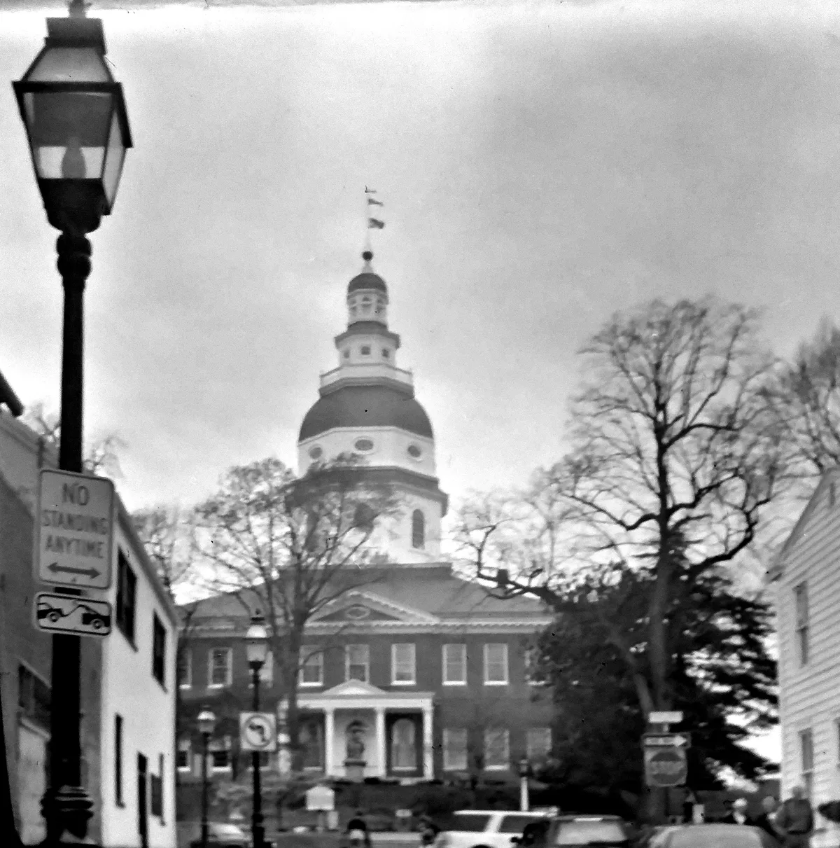 The Maryland State House rises at the end of a narrow street, its large domed cupola centered above the brick façade and framed by bare winter trees. A lamp post, parking signs, and a few parked cars line the foreground, giving the historic capitol a small-town street view.