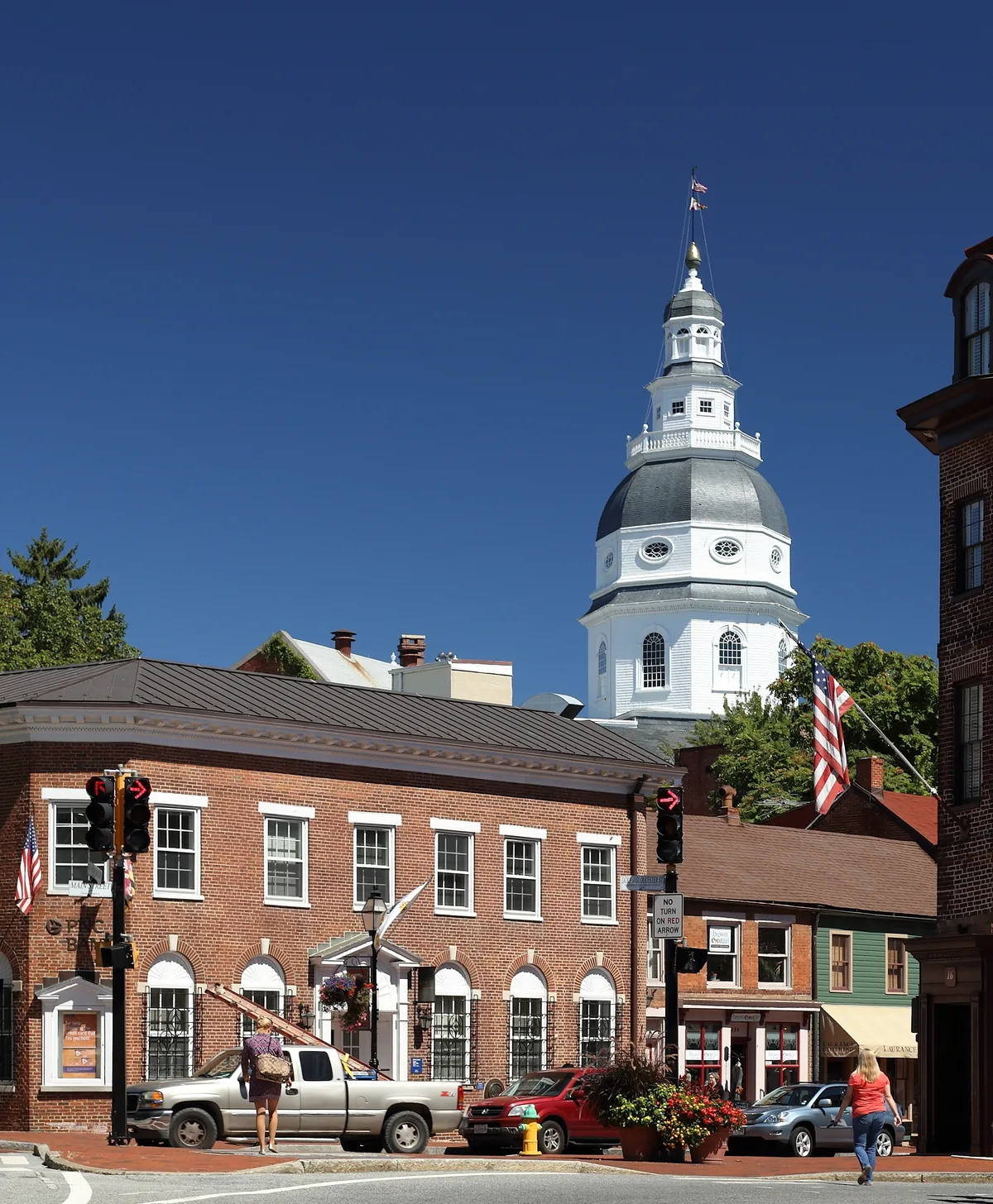 Maryland State House’s white wooden dome rises above a row of red-brick downtown Annapolis buildings, with American flags, parked cars, and pedestrians in the foreground. The clear blue sky and bright sunlight make the colonial architecture stand out sharply.