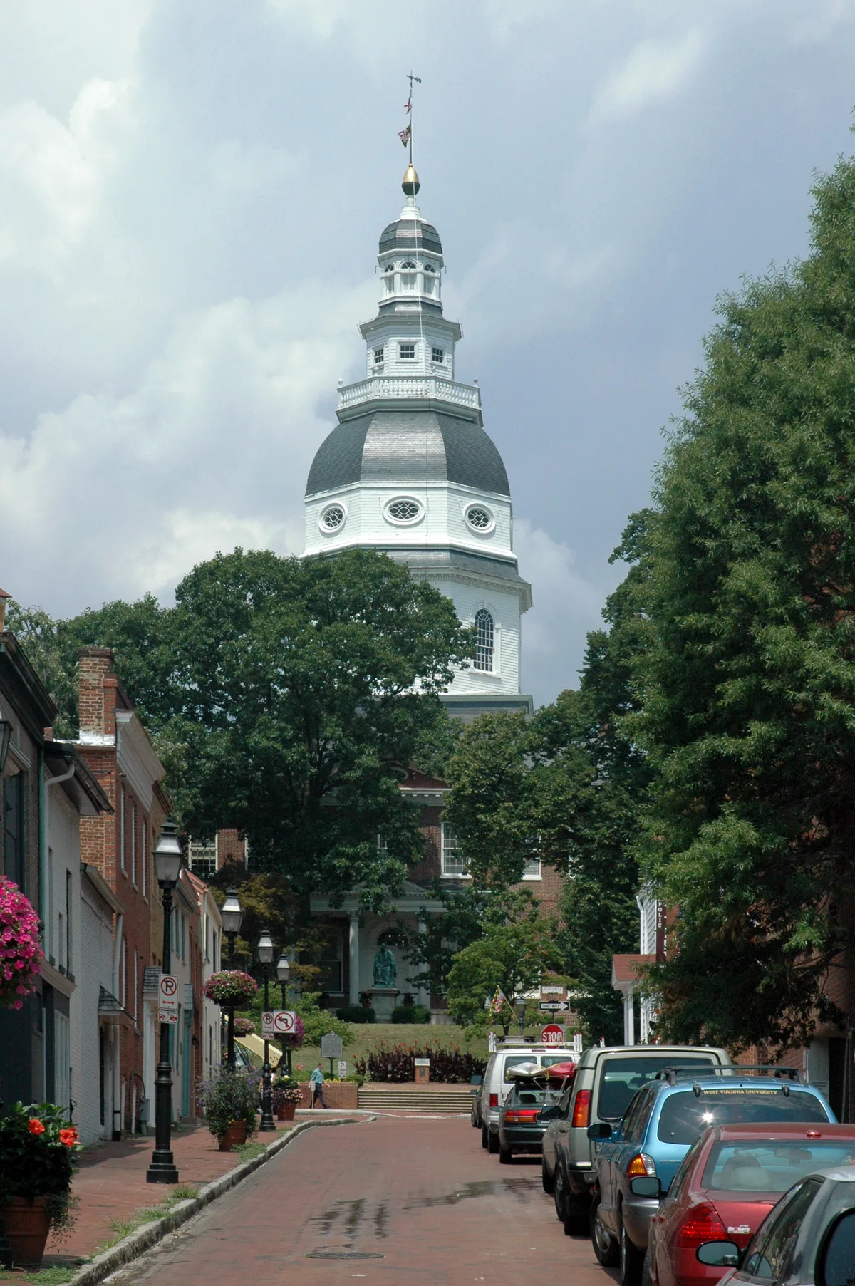 Looking up a brick-lined Francis Street toward the Maryland State House, with the tall white colonial dome rising above trees and historic row buildings. Parked cars, hanging flower baskets, and streetlamps frame the narrow street in soft daylight.