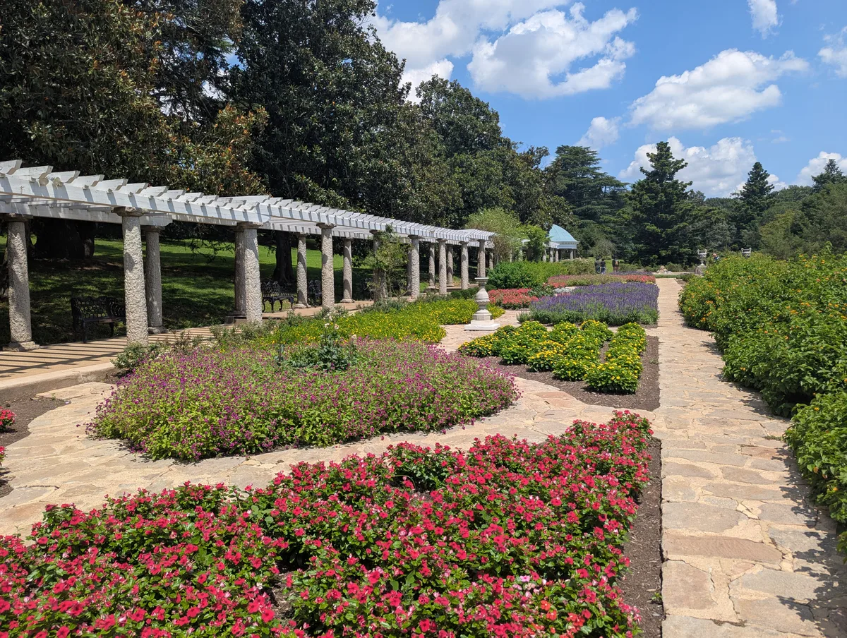 Sunlit Italian-style garden beds at Maymont stretch along stone walkways, with vivid pink, purple, yellow, and red flowers arranged in neat geometric plots. A long pergola of rough stone columns and white beams leads the eye toward the trees and blue sky beyond.
