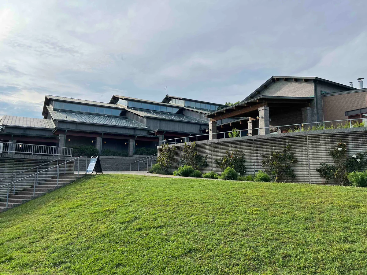 Robins Nature Center’s dark modern buildings step up a grassy slope, with wide concrete stairs on the left and a low planted retaining wall on the right. Soft morning light and a pale sky give the scene a calm, open feel.