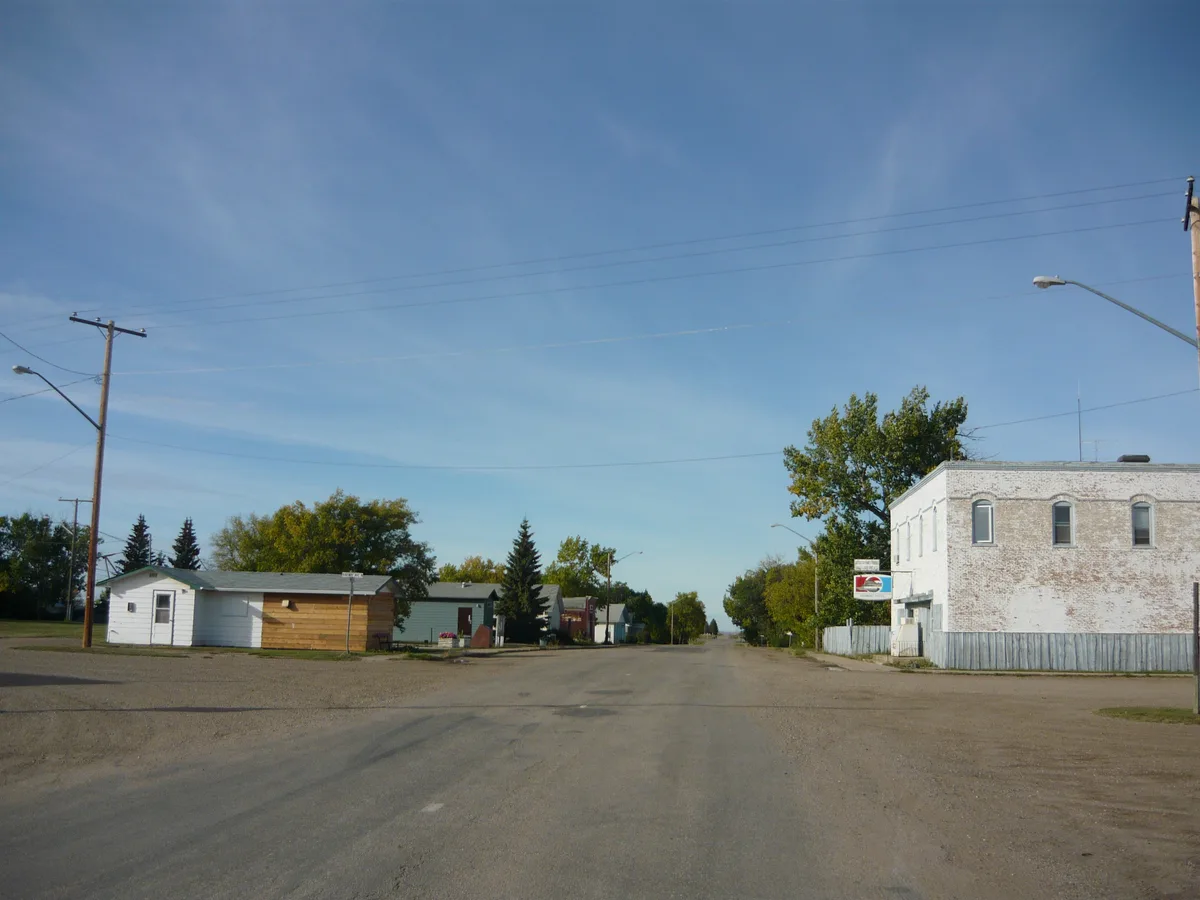 A quiet, wide street stretches into the distance under a clear blue morning sky, lined with small low-rise buildings, utility poles, and leafy trees. On the right, a weathered white brick building with a faded sign and fenced side yard anchors the foreground.
