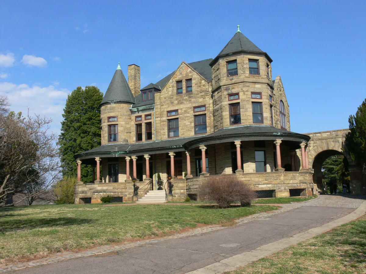 Maymont’s Gilded Age mansion stands in bright morning light, with tan stone walls, round corner turrets, and a wraparound porch topped by dark conical roofs. A curved walkway leads across the lawn toward the entrance, with bare trees and a clear blue sky framing the house.