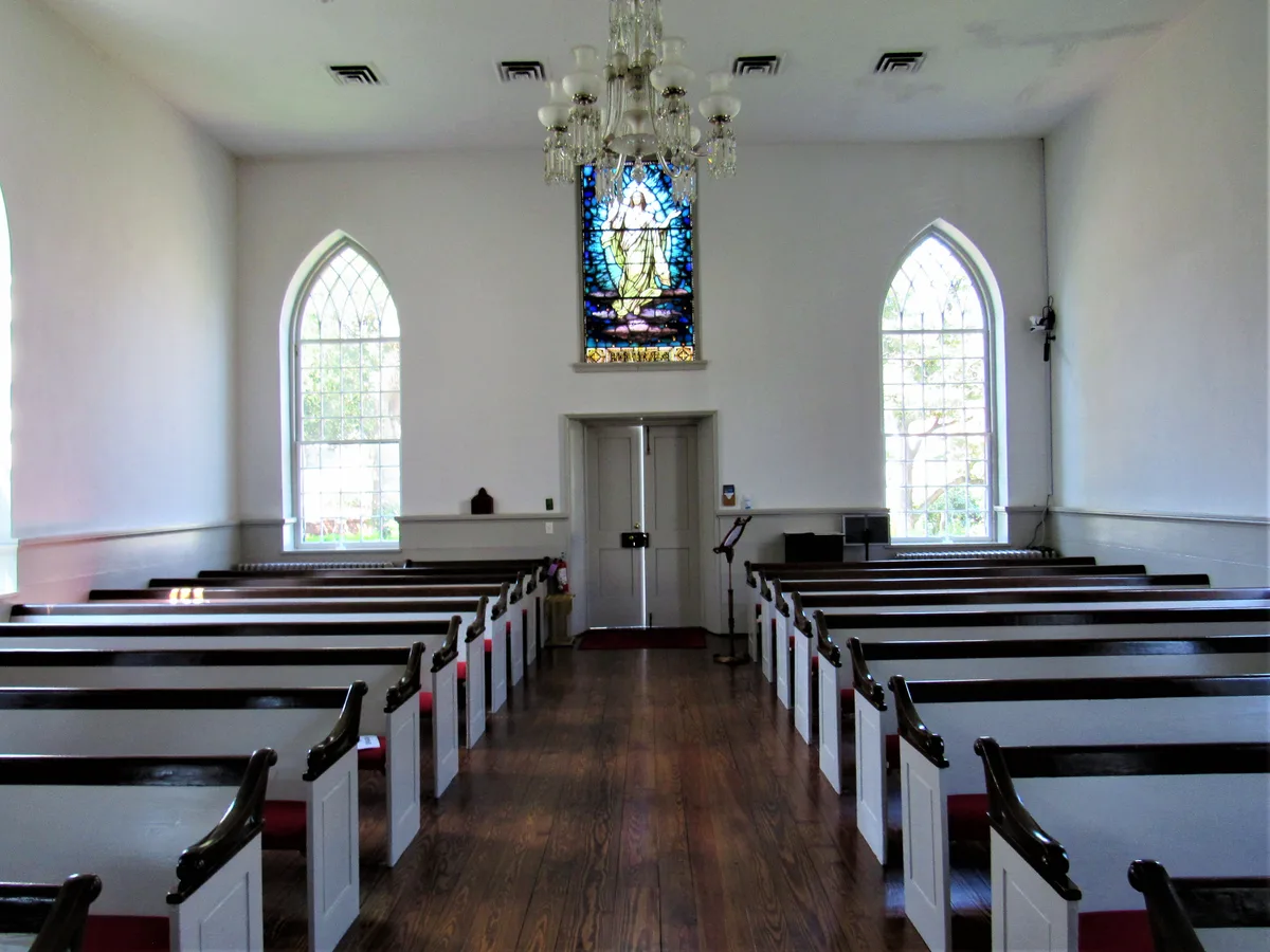 Bright, simple church interior with rows of white pews and dark wooden seats leading to a centered stained-glass window above the front doors. Tall pointed windows and a small chandelier brighten the white walls and polished wood floor with soft morning light.