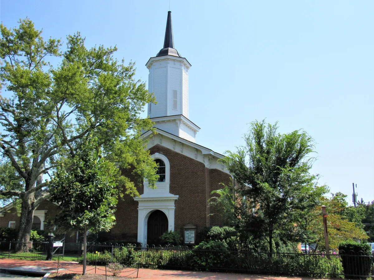 A white-steepled brick church rises behind leafy trees and a black iron fence, with an arched entrance and tall, narrow windows catching bright morning light. The scene feels quiet and settled, framed by mature greenery and a red-brick sidewalk in front.