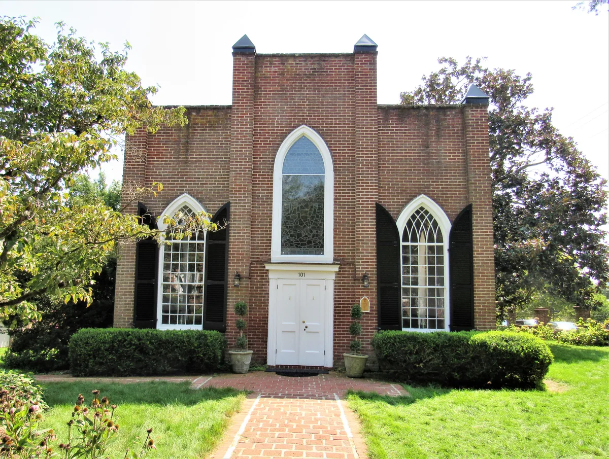 A small brick church stands front and center, with tall pointed-arch windows, black shutters, and a white double door reached by a brick path. Trimmed hedges, potted topiaries, and leafy trees frame the sunlit façade against a bright morning sky.