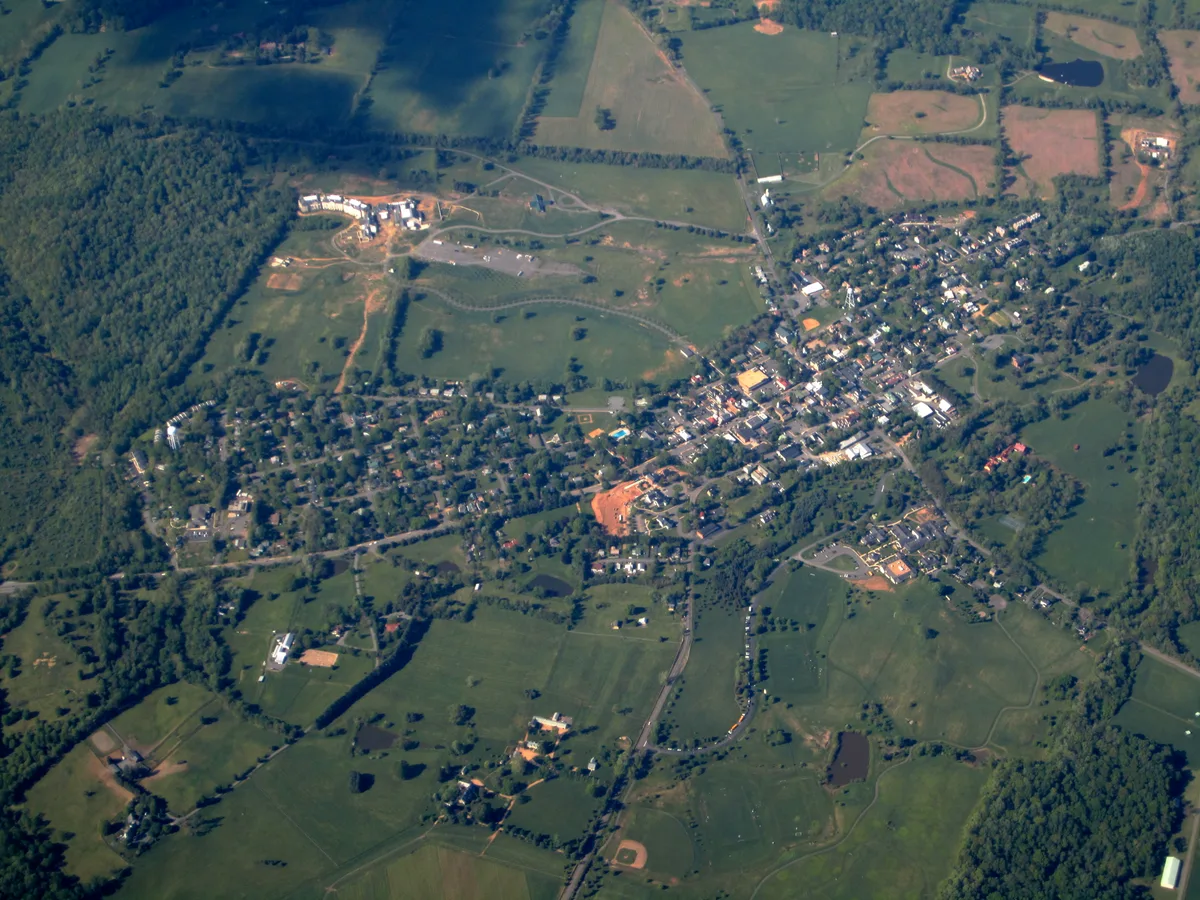 High aerial view of Middleburg set amid rolling green pastureland, with the town’s compact grid of streets and clustered rooftops centered among scattered estates and fields. Dark tree lines, winding roads, and a few reflective ponds frame the horse-country landscape on a clear morning.