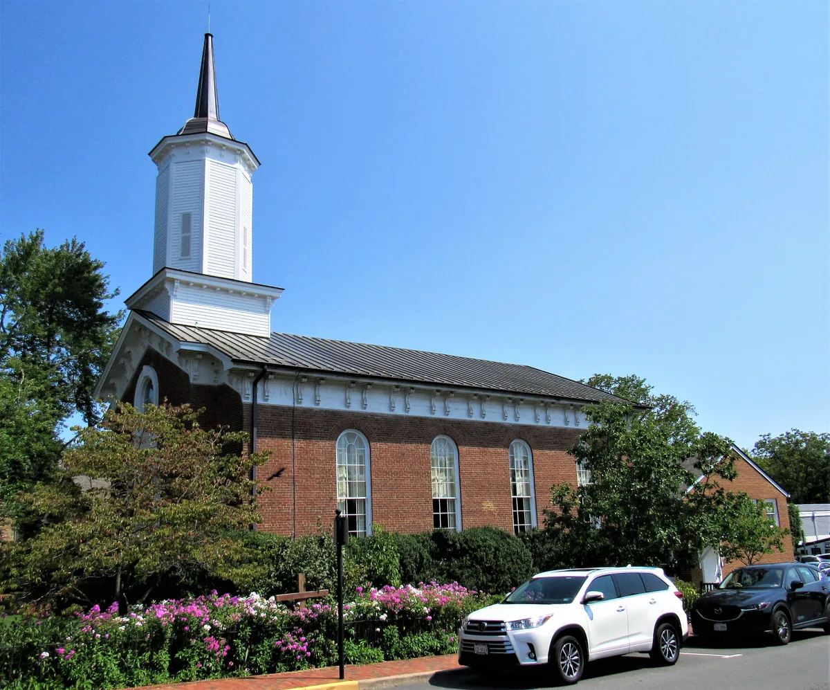 A red-brick church with tall arched windows and a white steeple rises above a street lined with parked cars, shrubs, and blooming pink flowers under a clear blue morning sky. The composition emphasizes the church’s classic small-town architecture and its leafy, well-kept setting.