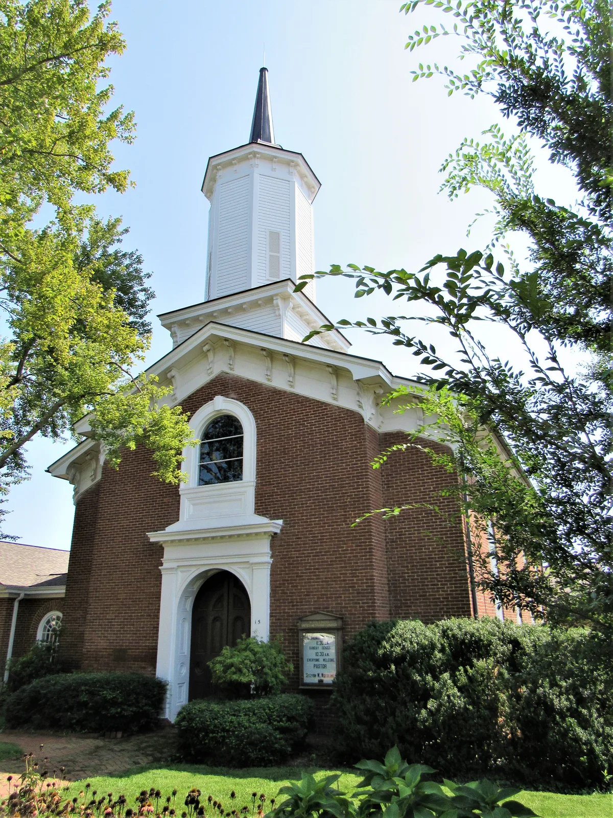 A brick church with white trim and a tall steeple rises above manicured shrubs and a green lawn, framed by leafy branches against a bright morning sky. The low angle emphasizes the entrance arch, tall arched window, and crisp Federal-style details.