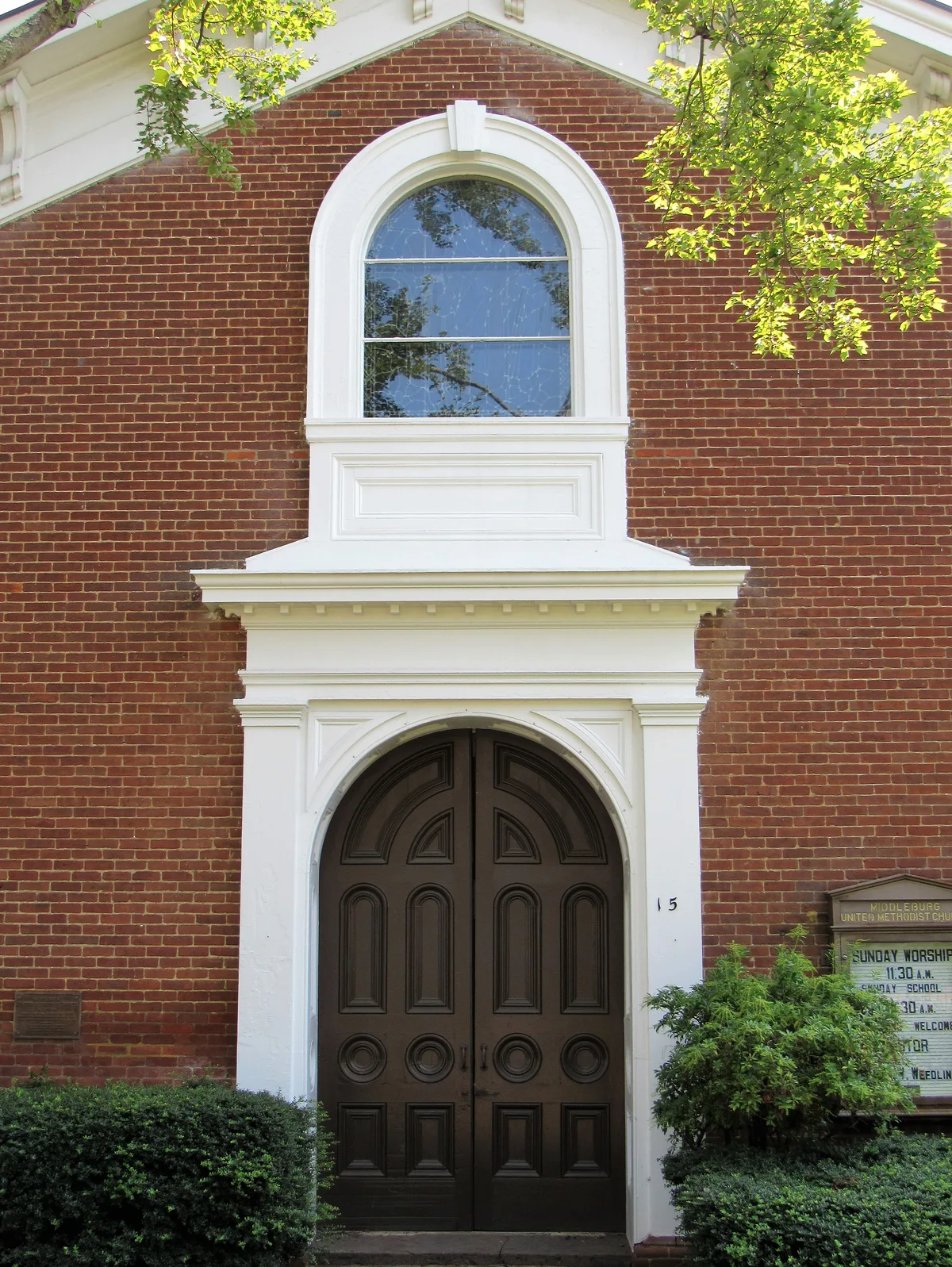 Close-up of a red-brick church facade in Middleburg with a white arched doorway, dark double doors, and a matching arched window above. Morning light filters through green leaves at the top edge, with trimmed shrubs and a small sign framing the entrance.