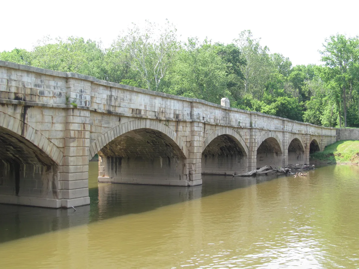 Seven white stone arches of the Monocacy Aqueduct stretch across the calm, greenish Monocacy River, with the long bridge curving gently through a leafy summer landscape. Soft morning light brightens the quarry stone and the water below.