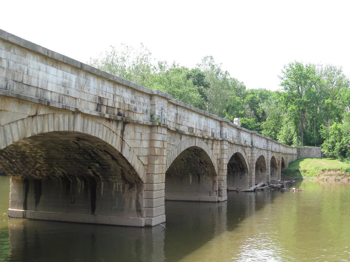 The Monocacy Aqueduct’s white stone arches stretch across the calm river, with dark water reflecting the underside of the structure and green trees lining the bank. Soft morning light highlights the weathered masonry and the long curve of the bridge.