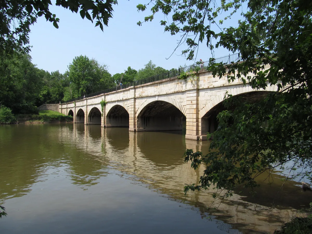 The white stone Monocacy Aqueduct stretches across the calm river in a wide view, its seven arches reflected in the water. Green trees frame the bridge, with leafy branches hanging into the foreground under a clear morning sky.