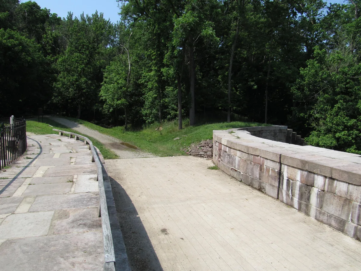 Stone canal walls and a broad wooden towpath curve across the Monocacy Aqueduct, with a black iron railing on the left and dense summer trees filling the background. Bright morning light highlights the pale quarry stone and the quiet, forested setting above the river crossing.