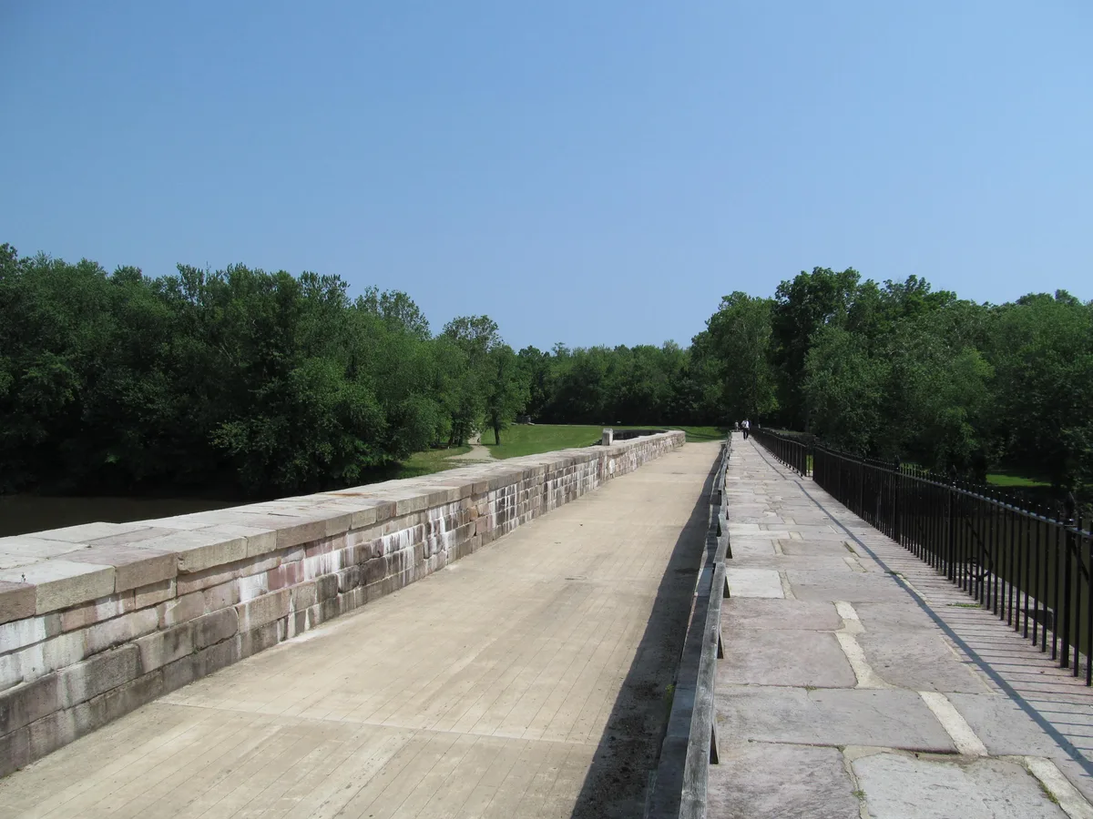 Wide view along the top of the Monocacy Aqueduct, with a pale stone canal bed stretching into the distance beside a narrow walkway and black iron railing. Dense green trees line both sides under a clear blue morning sky.