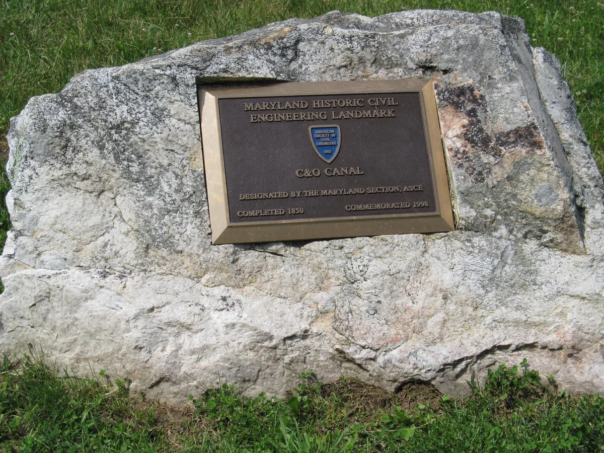 A close-up of a large gray boulder in grass with a bronze Maryland Historic Civil Engineering Landmark plaque mounted on it for the C&O Canal. The plaque’s dark metal surface and raised lettering contrast with the rough stone and green lawn.
