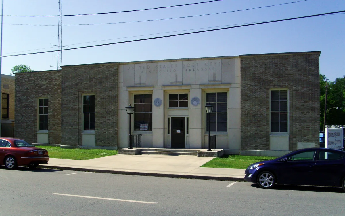 Sunlit, low-rise neoclassical post office with a flat roof, pale stone center, and brick wings, fronted by a simple concrete walk, black lamp posts, and parked cars along the street. The façade has tall rectangular windows and faint lettering above the entrance reading “United States Post Office Monticello Arkansas.”
