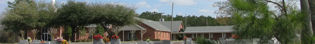 Wide banner view of a low brick building complex framed by trees and landscaped planters, with flower baskets adding bright color in the foreground. The scene is lit by soft morning sun under a blue sky with scattered clouds.