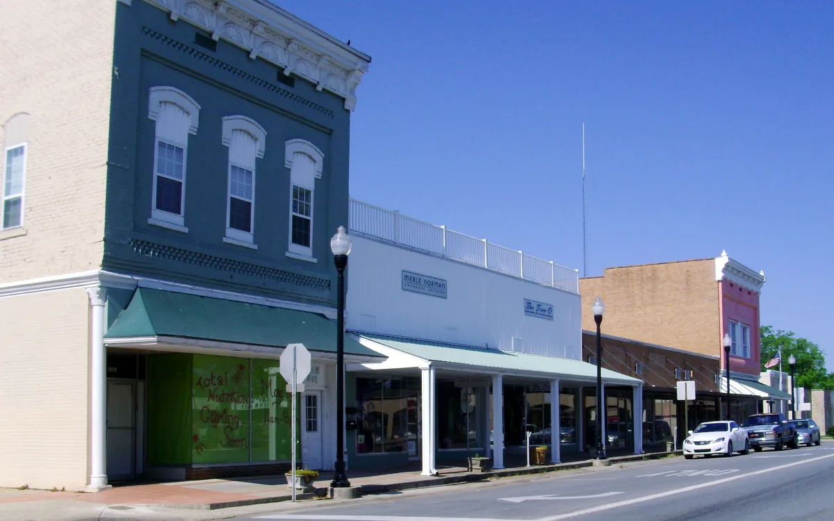 A row of small downtown storefronts lines a quiet street under a clear blue morning sky, with painted brick facades, awnings, and old-fashioned lamp posts. Parked cars and a few shop signs add to the historic small-town feel.