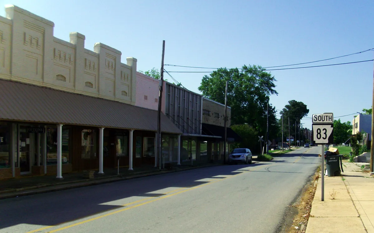 A quiet downtown street in bright morning light, lined with low historic storefronts and a decorative parapet along the left side. A South 83 highway sign, parked cars, overhead utility lines, and leafy trees lead the eye toward the distance under a clear blue sky.