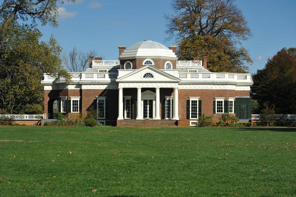 Monticello’s west facade stands centered behind a broad green lawn, with its white-columned portico, brick walls, and distinctive dome sharply lit in morning sun. Autumn trees frame the neoclassical estate against a clear blue sky.