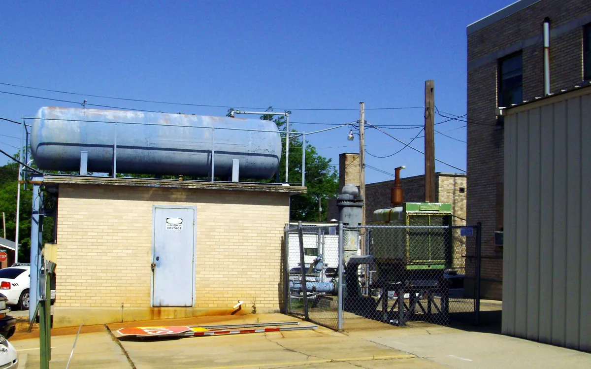 Industrial utility buildings and equipment fill the frame under a clear blue morning sky, with a large horizontal metal tank atop a small brick structure marked “High Voltage” and chain-link fencing around pipes and machinery. Utility poles, overhead wires, and a taller brick building at right give the scene a cramped, back-lot feel.