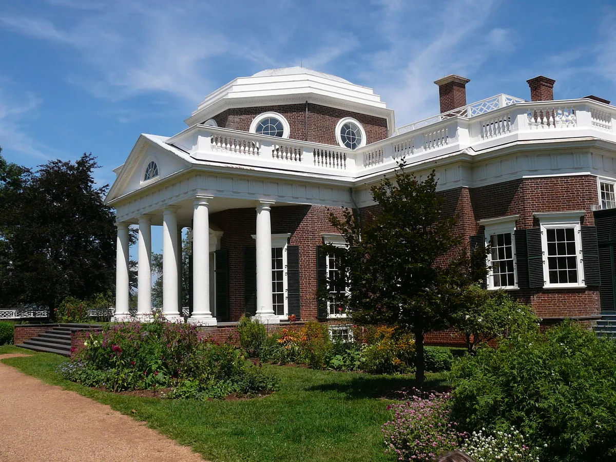Monticello’s brick west facade fills the frame, with its white columned portico, circular windows, and octagonal dome set against a bright blue morning sky. In front, manicured garden beds, a clipped lawn, and a gravel path soften the formal neoclassical architecture.