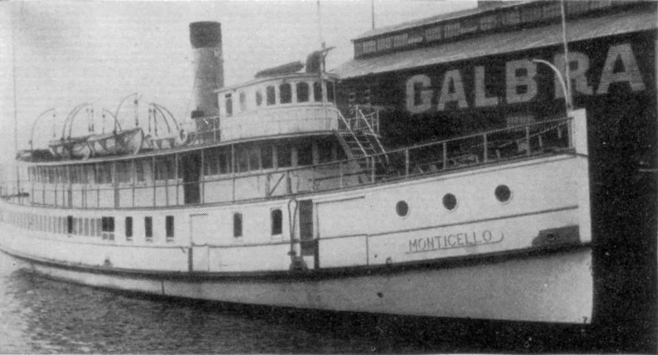 A black-and-white side view of the steamboat Monticello moored beside a large dock, with its white hull, row of portholes, upper decks, and lifeboats clearly visible. The boat’s name is painted near the bow, and the large “GALBRA” sign fills the background.