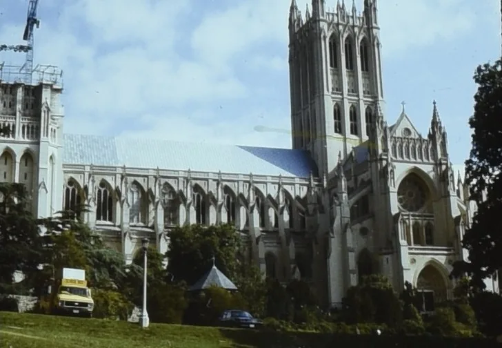 Washington National Cathedral rises above a grassy slope, with its tall Gothic tower, pointed arches, and flying buttresses framed against a bright blue sky. Trees and a few parked cars sit at the base, emphasizing the cathedral’s scale and intricate stone façade.