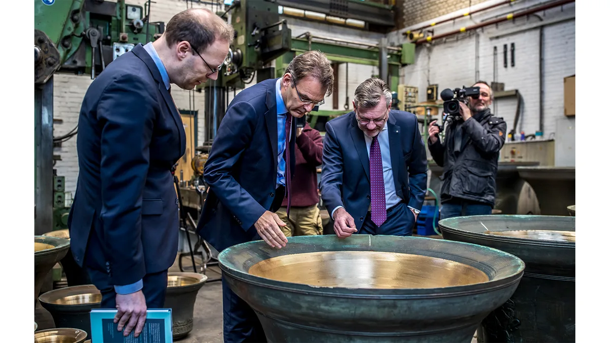 Three men in suits inspect a large bronze bell inside an industrial workshop, with a cameraman documenting the scene in the background. Overhead machinery, stacked bells, and white brick walls frame the close, documentary-style composition.