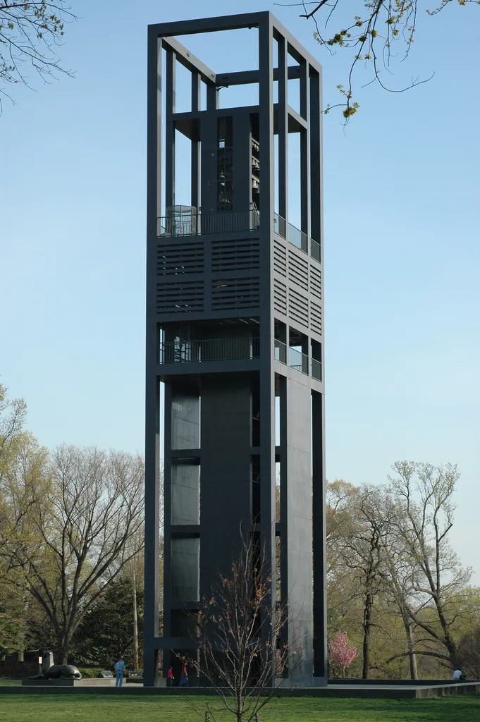 The Netherlands Carillon rises as a tall, open steel tower against a clear blue sky, with bare-limbed spring trees and a few people at its base on a grassy hill. The dark geometric frame stands in sharp contrast to the pale sky, giving the scene a clean, vertical composition.