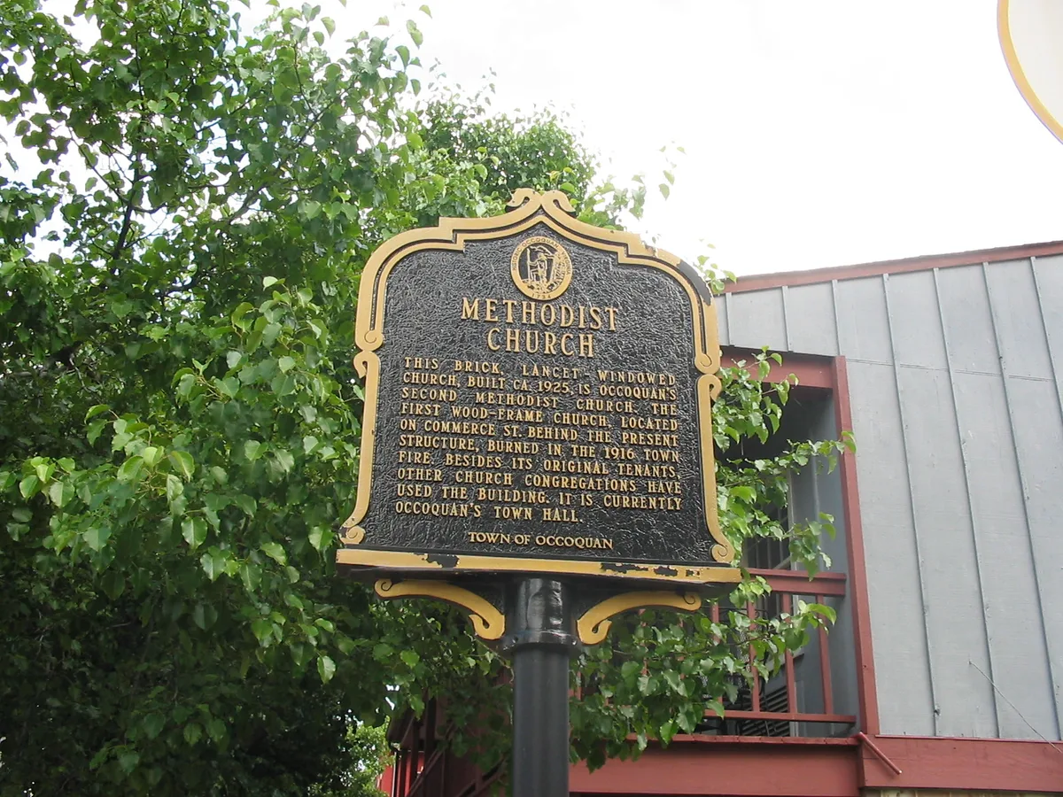 An ornate black-and-gold historic marker for Occoquan’s Methodist Church stands in the foreground, framed by leafy green trees. Behind it is a simple gray-and-red building with Victorian-style trim, under bright daylight.