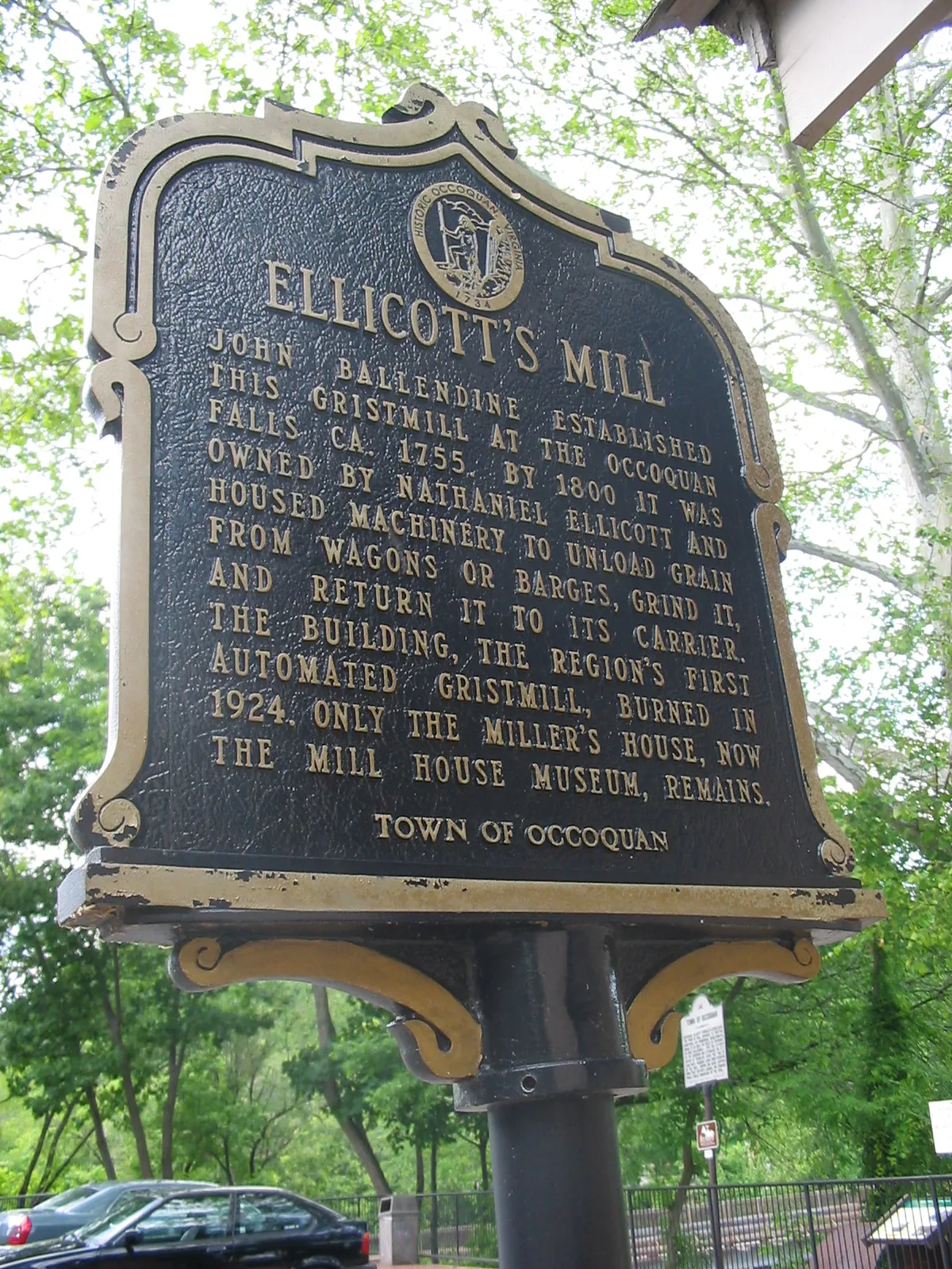 A large black-and-gold historic marker for Ellicott’s Mill fills the frame, with raised lettering and a decorative top against leafy green trees. The sign stands beside a road with parked cars and a glimpse of the riverfront park beyond.