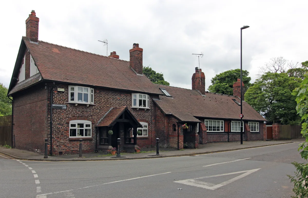 A quiet brick cottage with a steep red-tiled roof sits on a curved neighborhood street, its chimneys, white-trimmed windows, and small covered entry giving it a classic old-English feel. Overcast light softens the scene, with leafy trees and a lamppost framing the house against a pale sky.