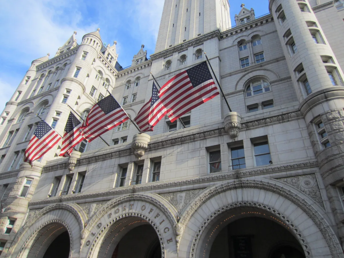 Low-angle view of the Old Post Office Pavilion’s pale stone facade, with ornate turrets, arched entrances, and a row of American flags fluttering across the front. Bright daylight highlights the building’s carved details against a blue sky.