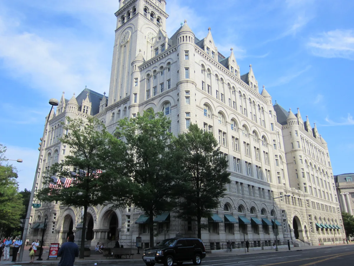 The Old Post Office Tower rises in pale stone with Gothic arches, turrets, and a tall clock tower against a bright blue sky. Trees partially cover the lower façade, where teal awnings, American flags, pedestrians, and a black SUV line the street below.