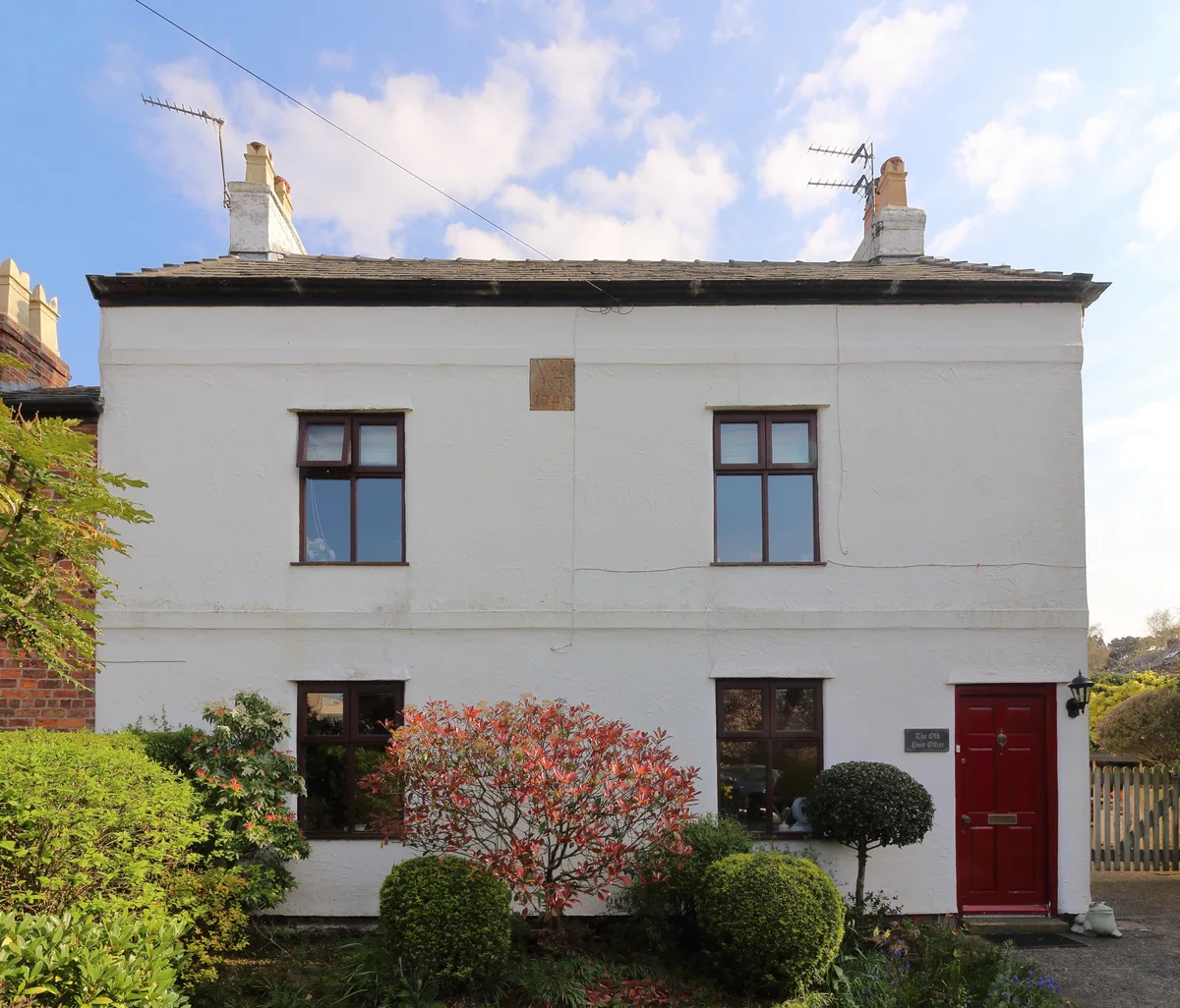 A small white stucco cottage with a red front door and dark-framed windows sits under a bright blue sky with scattered clouds. Trimmed shrubs and a red-leafed bush fill the front garden, giving the house a tidy, sunlit village feel.
