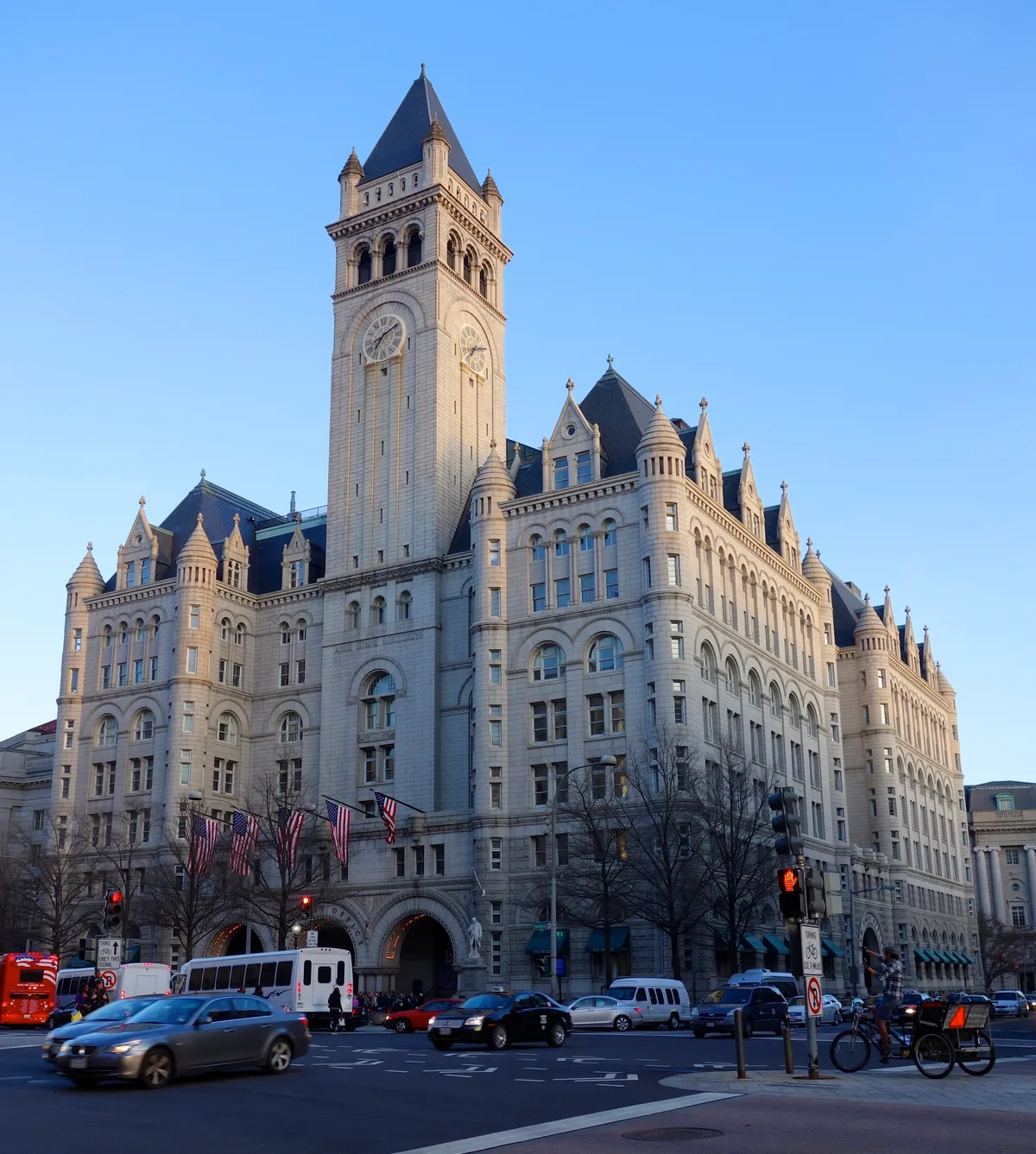 The Old Post Office Tower rises over a busy downtown Washington, DC intersection, its pale stone facade and clock tower catching warm golden-hour light against a clear blue sky. American flags hang above the arched entrances while cars, a bus, and pedestrians move through the street below.