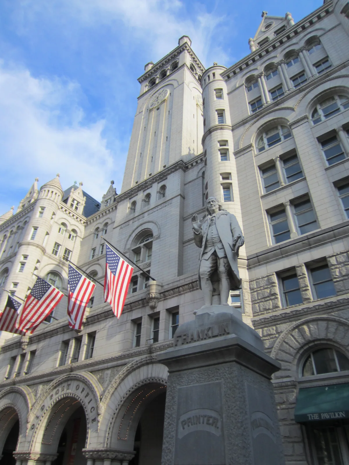 Low-angle view of the Old Post Office Pavilion’s pale stone facade, with its clock tower rising into a bright blue sky and Benjamin Franklin’s statue in the foreground. Three American flags hang above the arched entrance, and the ornate Romanesque details and round windows catch soft daylight.