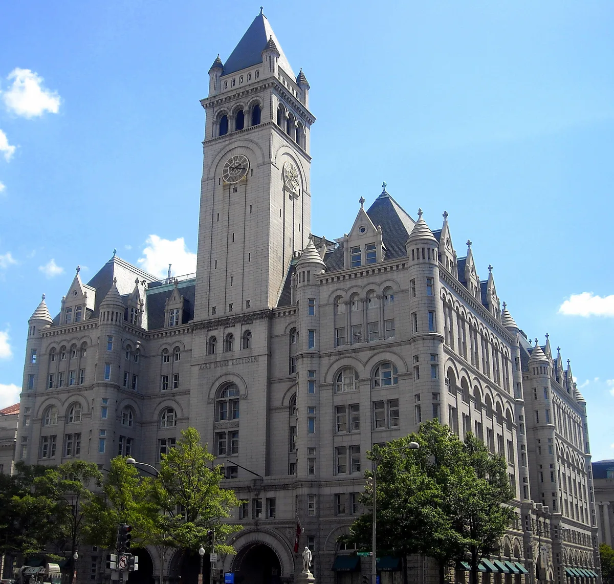 The Old Post Office Pavilion rises in pale stone with a tall central clock tower and steep slate roof, framed by ornate arches, turrets, and rows of arched windows. Bright blue sky and a few small clouds give the historic building a crisp, sunlit look.